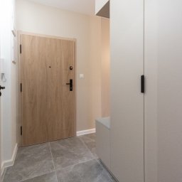 NIFU Jeremi Bombała - Nowoczesny przedpokój w neutralnych barwach z szarą marmurową podłogą, drewnianymi drzwiami wejściowymi z czarną klamką i beżową szafą na wymiar z siedziskiem.