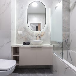 NIFU Jeremi Bombała - Nowoczesna łazienka z białymi marmurkowymi płytkami, owalnym podświetlanym lustrem, białą umywalką nablatową, beżową szafką pod umywalką z otwartymi półkami, białą wanną z szklanym parawanem...