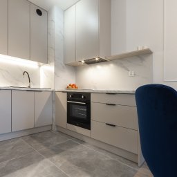 NIFU Jeremi Bombała - Nowoczesna kuchnia w odcieniach szarości z marmurowym blatem i backsplashiem, oświetleniem LED pod szafkami i płytkami podłogowymi imitującymi kamień, uzupełniona granatowymi krzesłami.