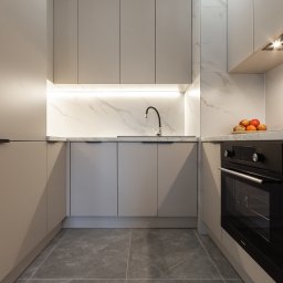 NIFU Jeremi Bombała - Nowoczesna kuchnia w odcieniach beżu z marmurowym blatem i podświetleniem LED, czarny piekarnik w zabudowie, owoce na blacie, szara podłoga z płytek.