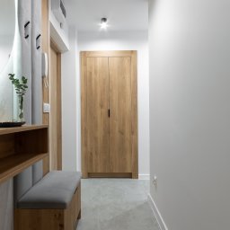 NIFU Jeremi Bombała - Nowoczesny przedpokój z drewnianą szafą wnękową z minimalistycznymi uchwytami, szarą podłogą z płytek, siedziskiem z szarą tapicerką i dekoracyjnym wazonem z zielonymi gałązkami na półce.