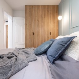 NIFU Jeremi Bombała - Sypialnia z drewnianą szafą na wymiar z czarnymi uchwytami, widok z perspektywy łóżka z szarą pościelą i dekoracyjnymi poduszkami w odcieniach niebieskiego i bieli, oświetlona wiszącą lampą kulą.