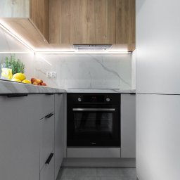 NIFU Jeremi Bombała - Nowoczesna kuchnia w odcieniach szarości i drewna, z czarnym piekarnikiem Amica, blatem z imitacji marmuru, podświetleniem LED i dekoracyjnym szkłem z cytrynami i ziołami.