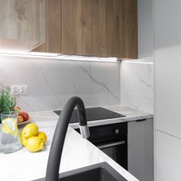 NIFU Jeremi Bombała - Nowoczesna kuchnia z drewnianymi szafkami górnymi, marmurowym blatem i backsplashiem, czarnym zlewem i baterią, z owocami i ziołami jako elementem dekoracyjnym, oświetleniem LED pod szafkami...