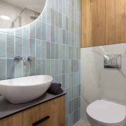 NIFU Jeremi Bombała - Nowoczesna łazienka z umywalką nablatową, armaturą chromowaną, pionowymi, błękitnymi płytkami i drewnianą szafką pod umywalką, w tle wisząca toaleta i drewniana szafka.