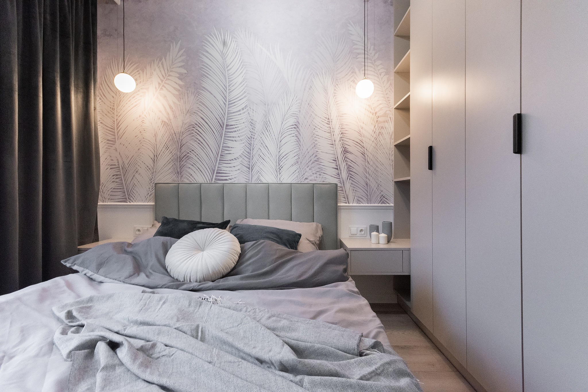 Nowoczesna sypialnia z tapetą w motywie liści palmowych, szarą tapicerowaną ramą łóżka, okrągłą białą poduszką dekoracyjną i wysoką, białą szafą w zabudowie z czarnymi uchwytami.