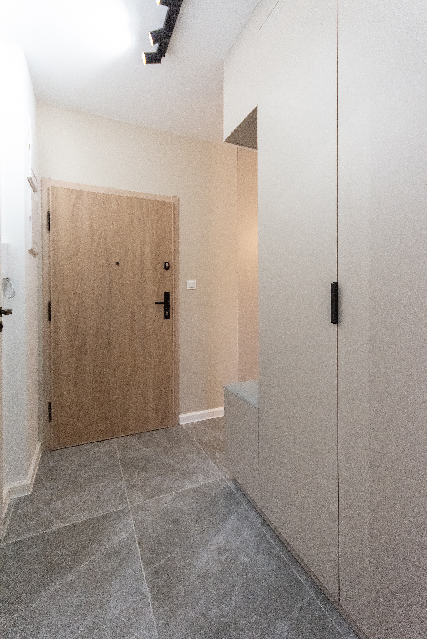 Nowoczesny przedpokój w neutralnych barwach z szarą marmurową podłogą, drewnianymi drzwiami wejściowymi z czarną klamką i beżową szafą na wymiar z siedziskiem.