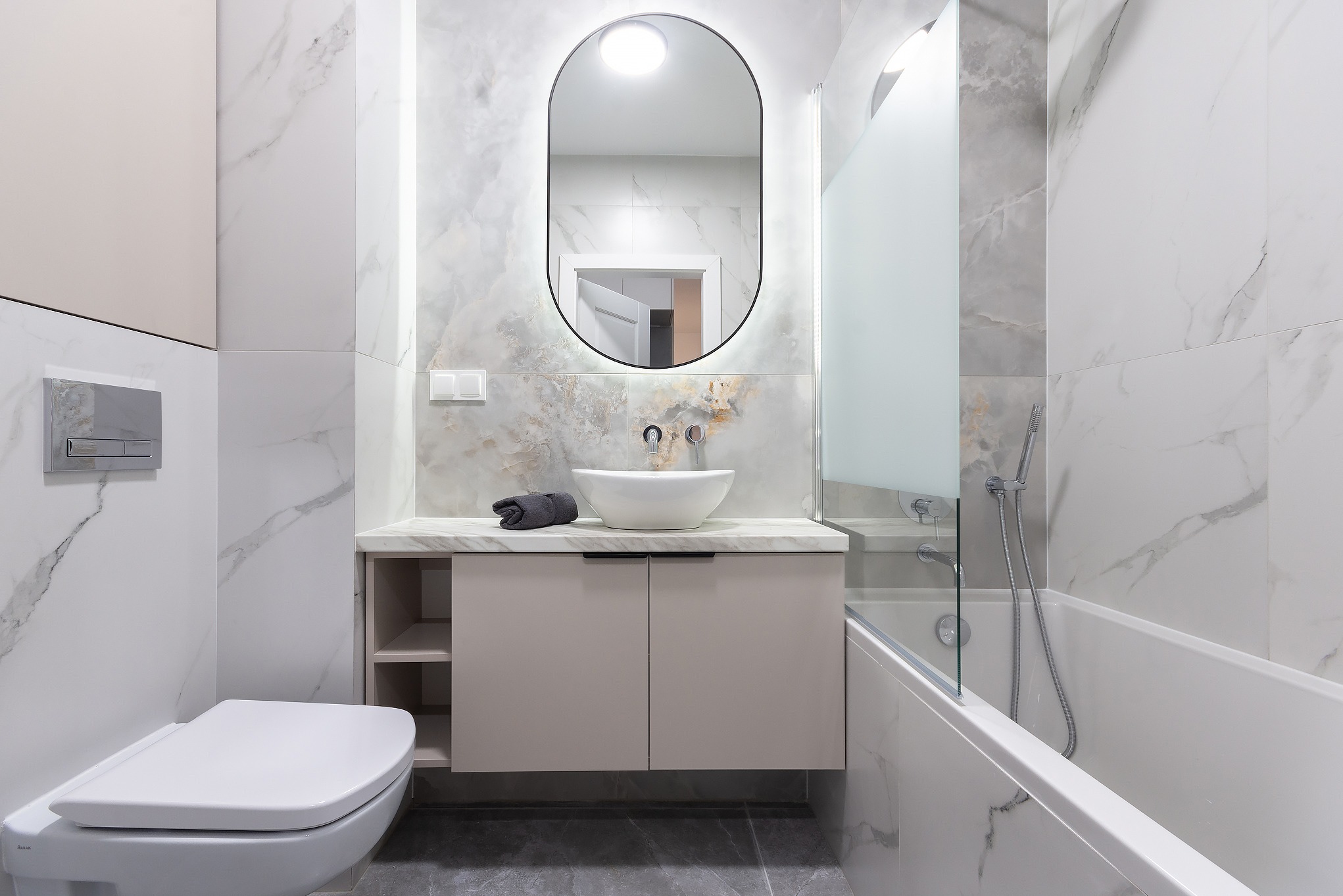 Nowoczesna łazienka z białymi marmurkowymi płytkami, owalnym podświetlanym lustrem, białą umywalką nablatową, beżową szafką pod umywalką z otwartymi półkami, białą wanną z szklanym parawanem...