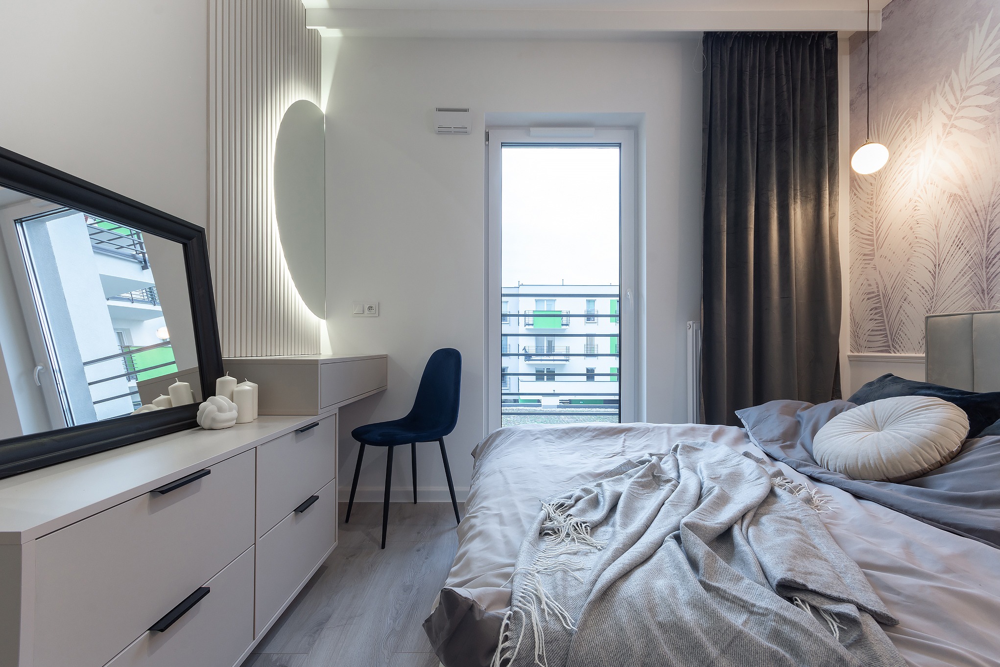Nowoczesna sypialnia w jasnych barwach z łóżkiem, komodą z lustrem w czarnej ramie, krzesłem i oknem balkonowym z widokiem na budynek mieszkalny.