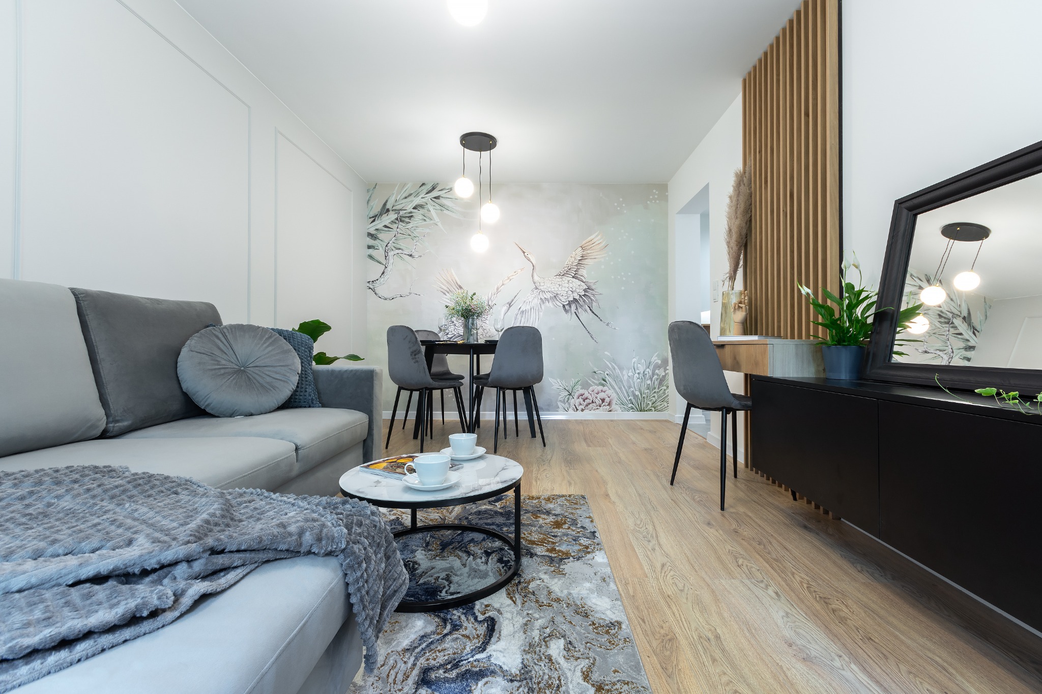 Nowoczesne wnętrze salonu z szarą sofą, stolikiem kawowym z marmurowym blatem, drewnianą podłogą, ścianą z motywem żurawi i lampą wiszącą z trzema kulami.