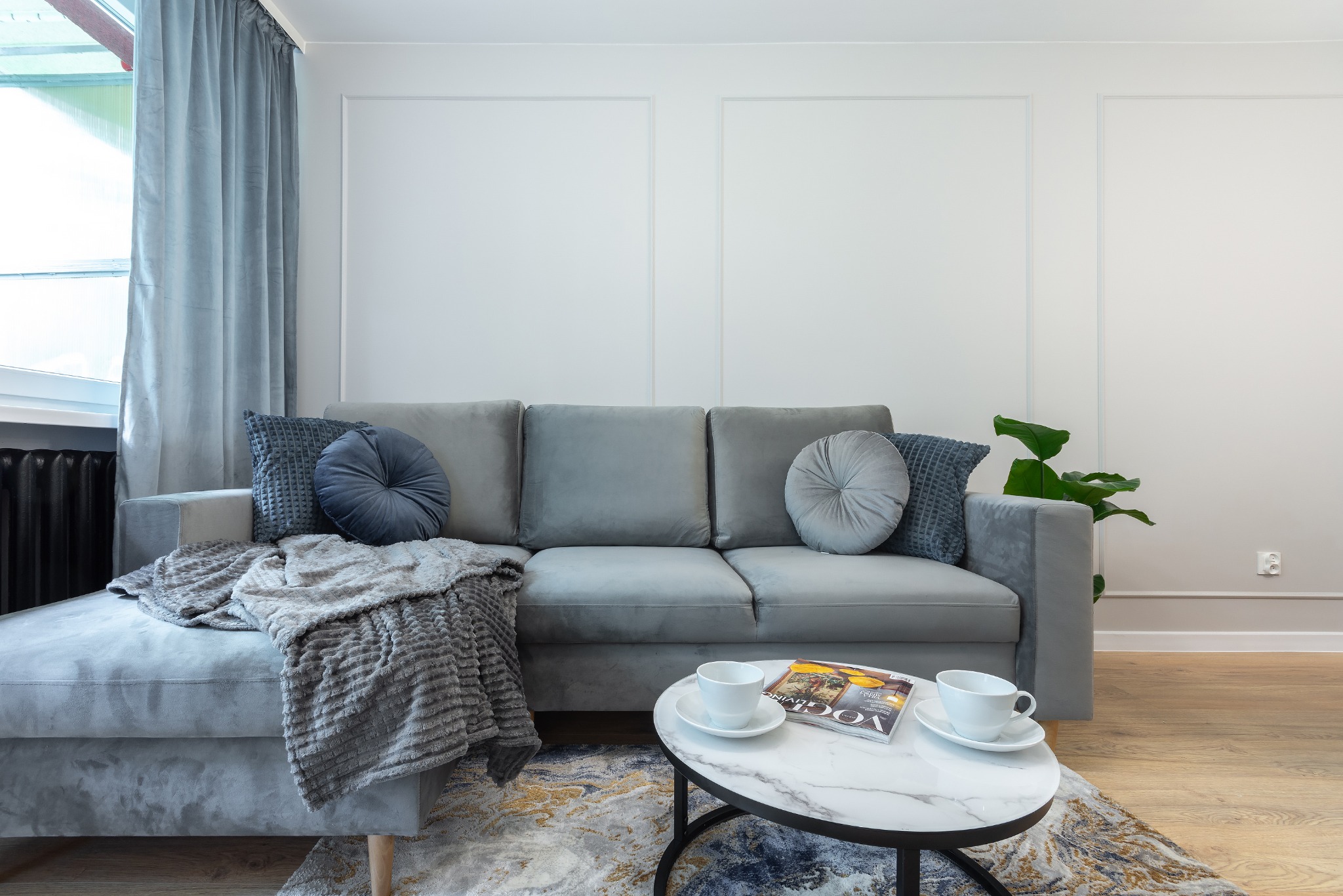 Szary narożnik z poduszkami dekoracyjnymi i pledem, stolik kawowy z marmurowym blatem, dwie filiżanki i magazyn, na podłodze dywan wzorzysty, ściana z białymi panelami dekoracyjnymi, zasłony w oknie.