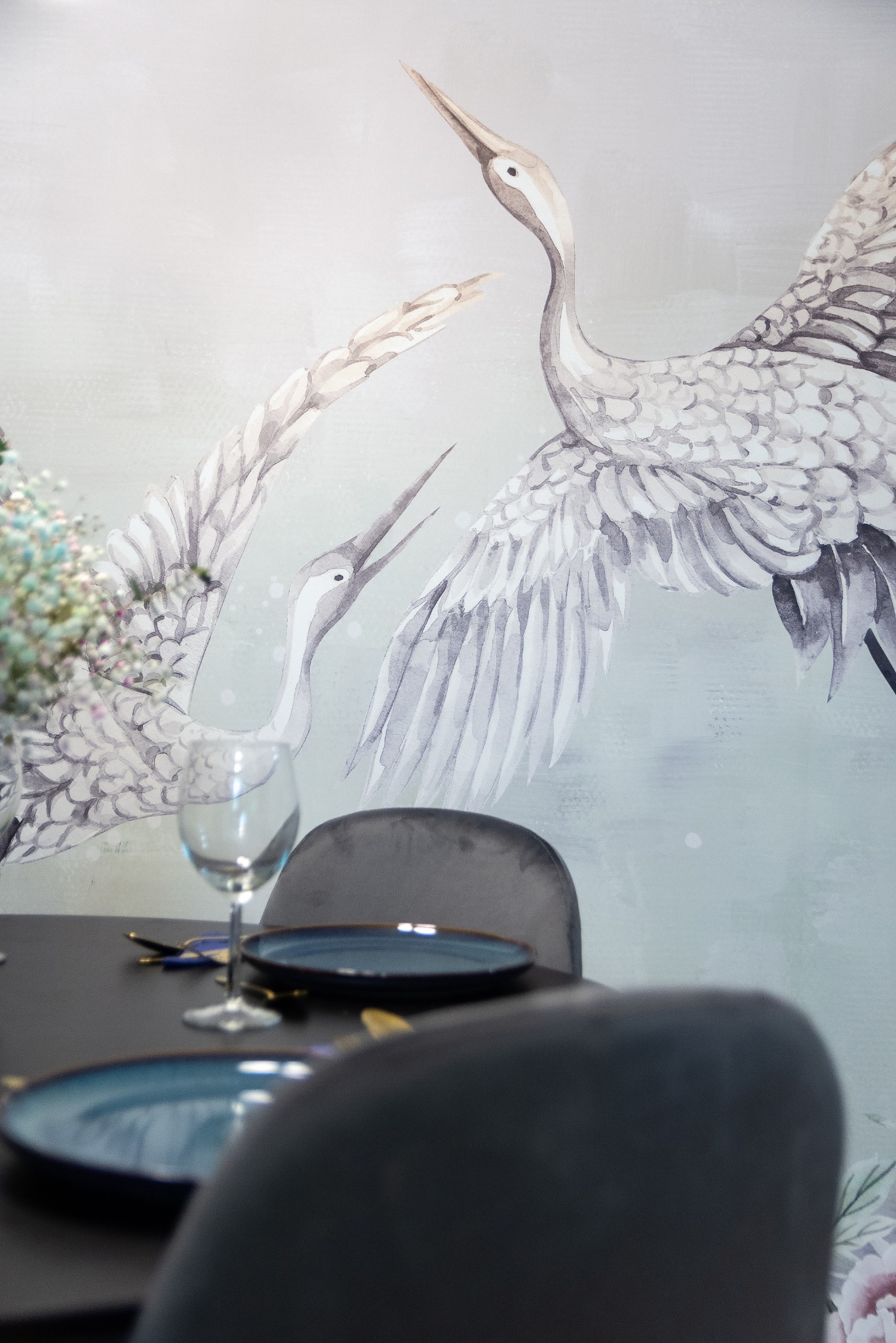 Stół z ciemnym blatem i nakryciem, na tle ściany z malowidłem dwóch żurawi w stylu akwareli, widoczne krzesła z welurową tapicerką i kwiaty w wazonie.
