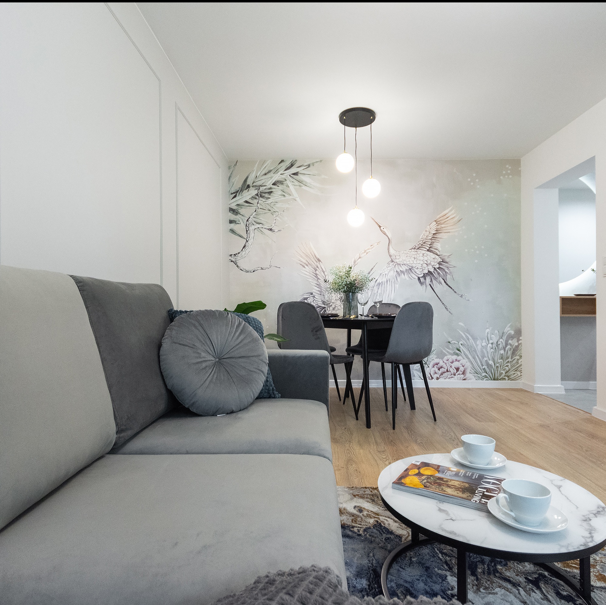 Jasne wnętrze salonu z elementami dekoracyjnymi: szary narożnik z okrągłą poduszką, okrągły stolik kawowy z marmurowym blatem i filiżankami, stół z krzesłami na tle ściany z motywem żurawi i roślin.