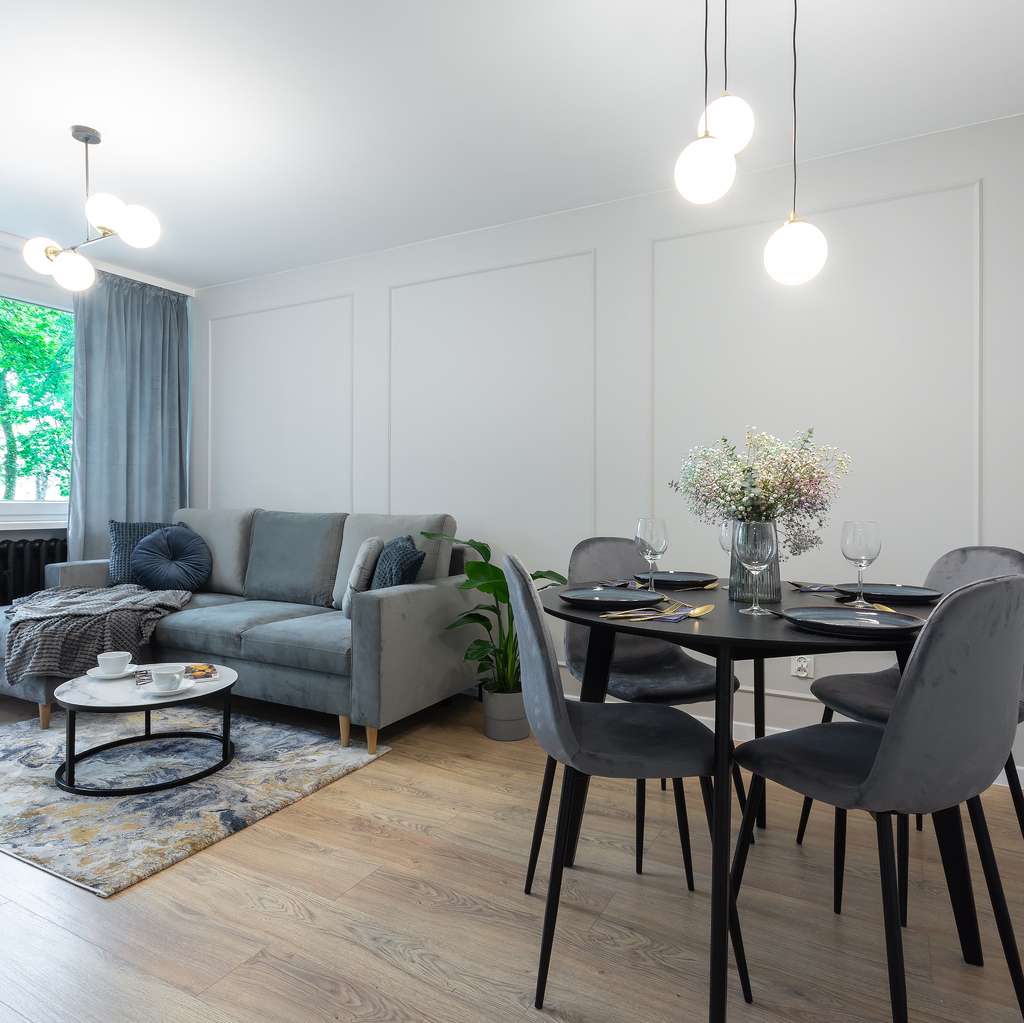 Nowoczesne wnętrze salonu z szarym narożnikiem, okrągłym stolikiem kawowym, okrągłym stołem jadalnym z szarymi krzesłami i dekoracyjną ścianą w ramkach.