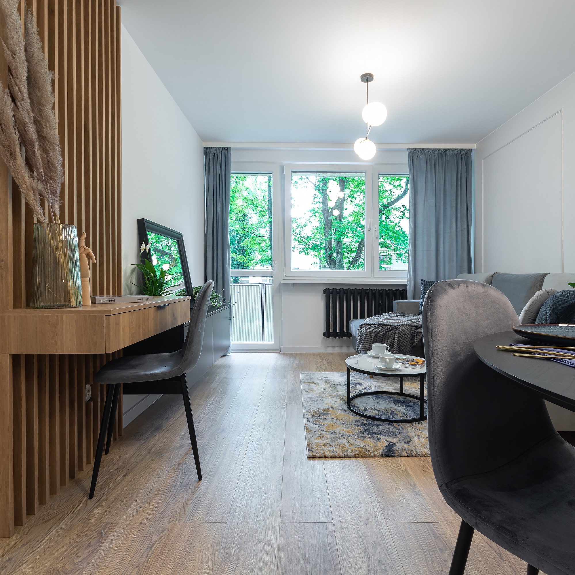 Jasne wnętrze pokoju z drewnianą podłogą, biurkiem z drewnianymi lamelami, szarą tapicerowaną krzesłem, szarą sofą, okrągłym stolikiem kawowym i dużym oknem z widokiem na zieleń