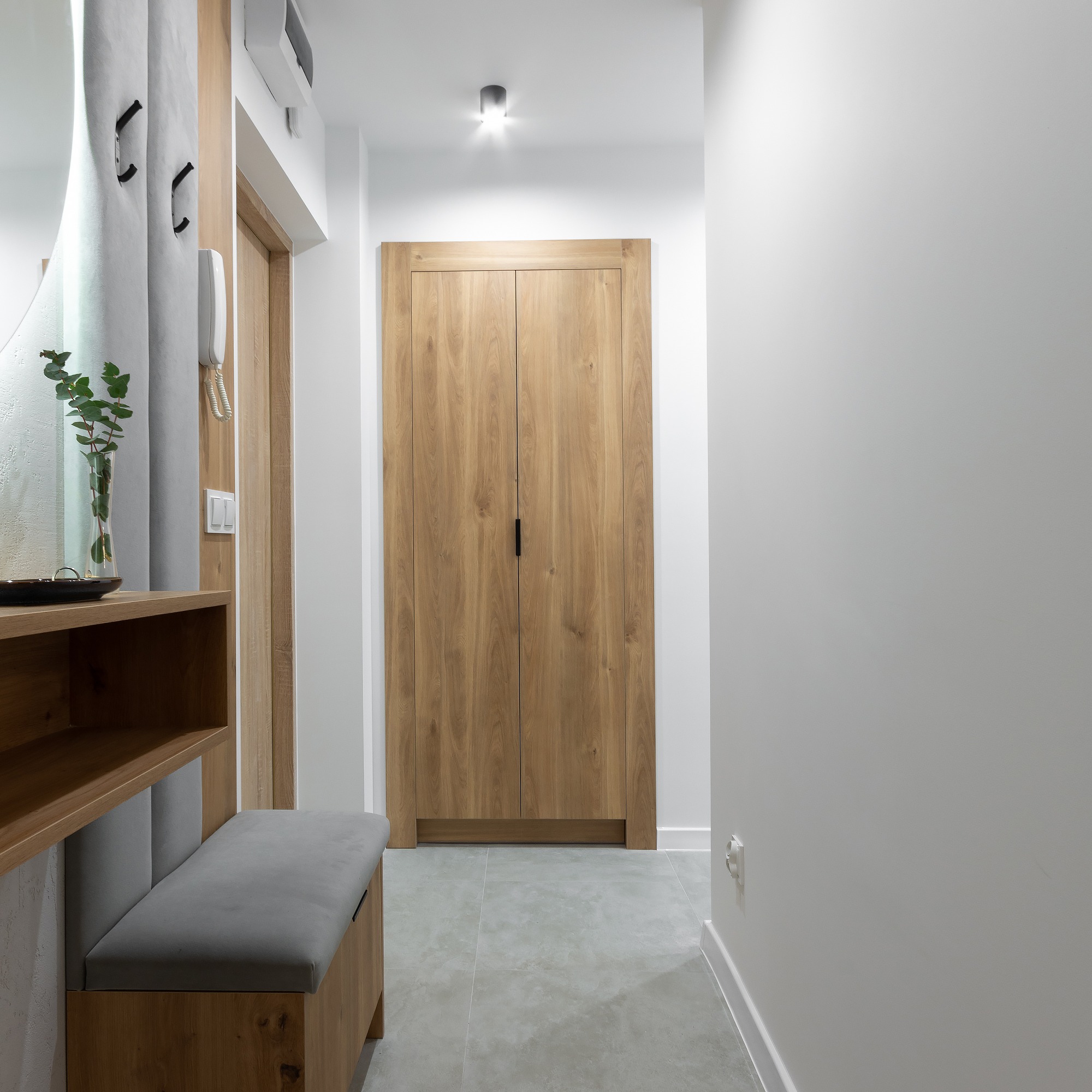 Nowoczesny przedpokój z drewnianą szafą wnękową z minimalistycznymi uchwytami, szarą podłogą z płytek, siedziskiem z szarą tapicerką i dekoracyjnym wazonem z zielonymi gałązkami na półce.