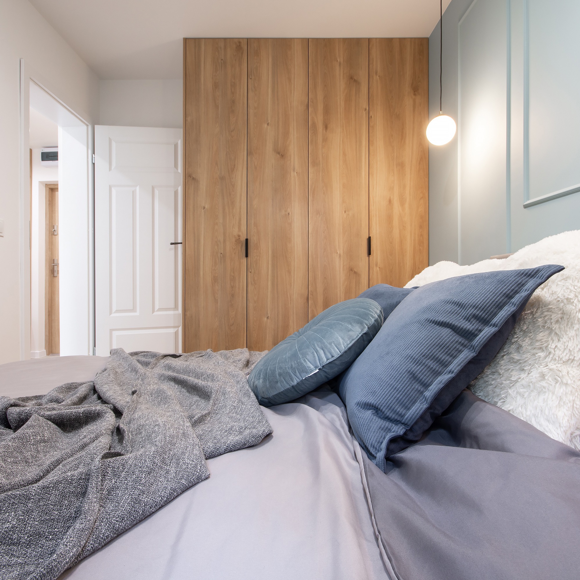 Sypialnia z drewnianą szafą na wymiar z czarnymi uchwytami, widok z perspektywy łóżka z szarą pościelą i dekoracyjnymi poduszkami w odcieniach niebieskiego i bieli, oświetlona wiszącą lampą kulą.