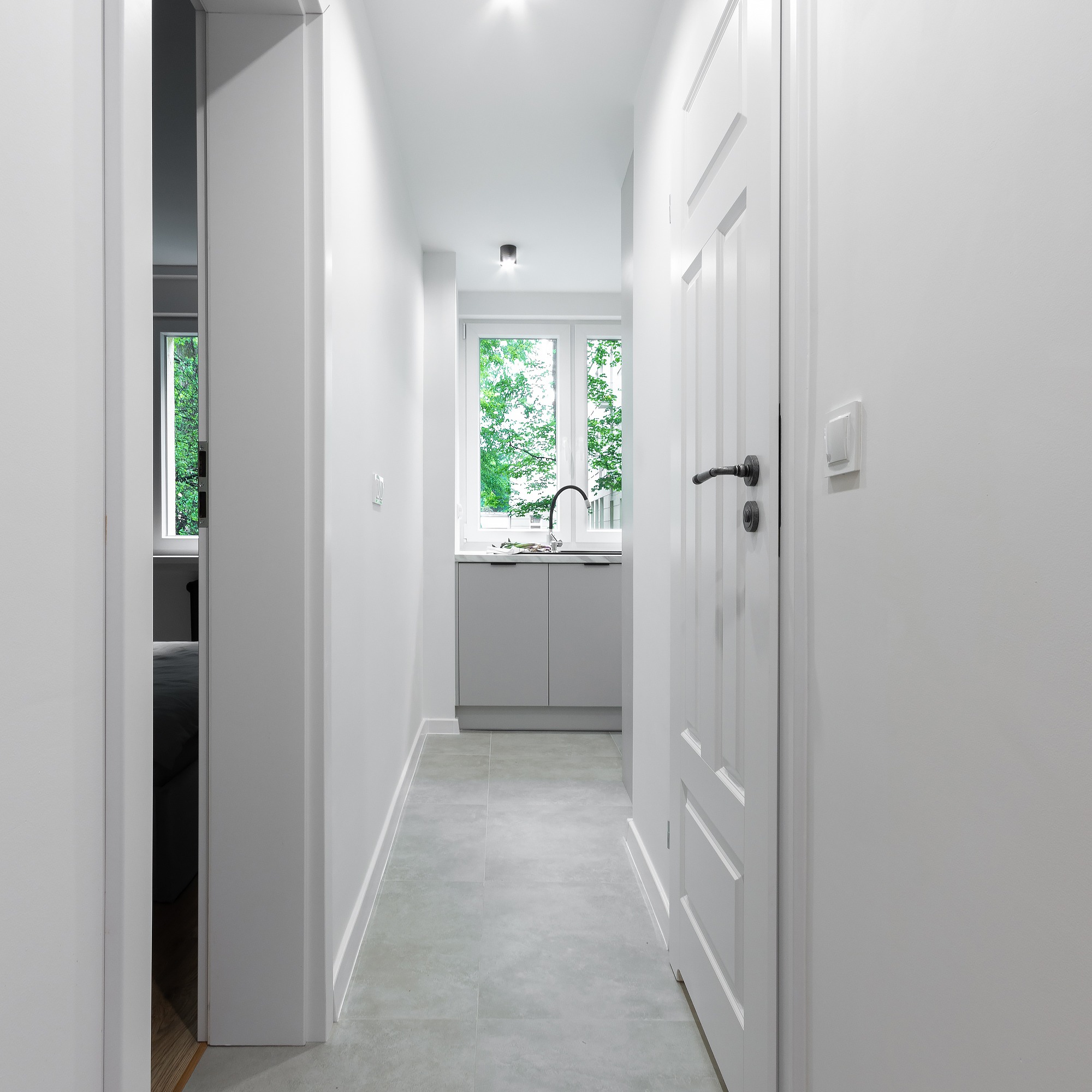 Wnętrze korytarza z szarymi płytkami podłogowymi, białymi ścianami i drzwiami, widok na aneks kuchenny z szarą zabudową i oknem z widokiem na zieleń.
