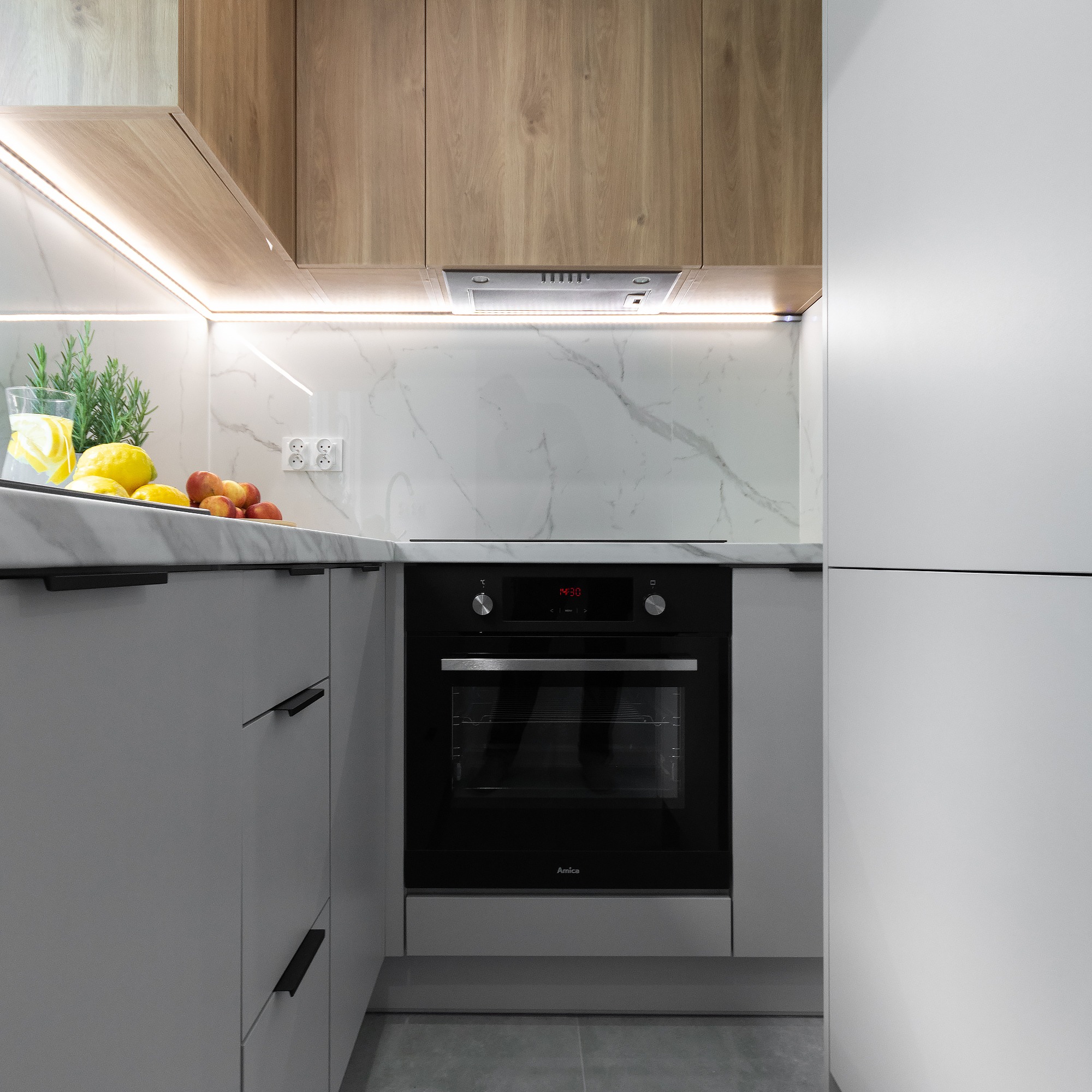 Nowoczesna kuchnia w odcieniach szarości i drewna, z czarnym piekarnikiem Amica, blatem z imitacji marmuru, podświetleniem LED i dekoracyjnym szkłem z cytrynami i ziołami.