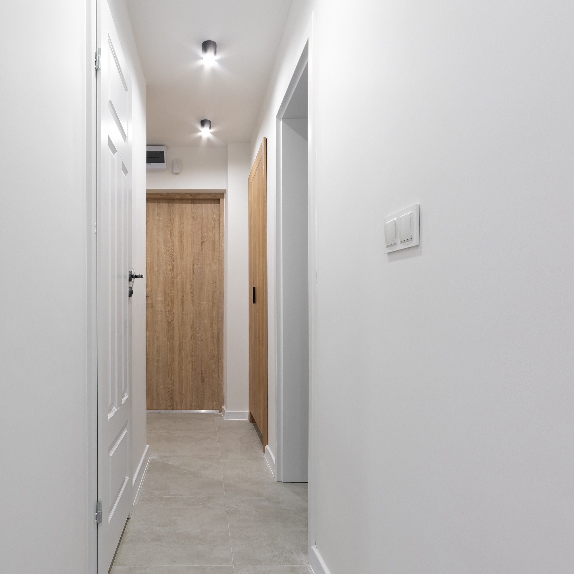 Nowoczesny korytarz z białymi ścianami, szarą podłogą z płytek, drewnianymi drzwiami i minimalistycznym oświetleniem sufitowym.
