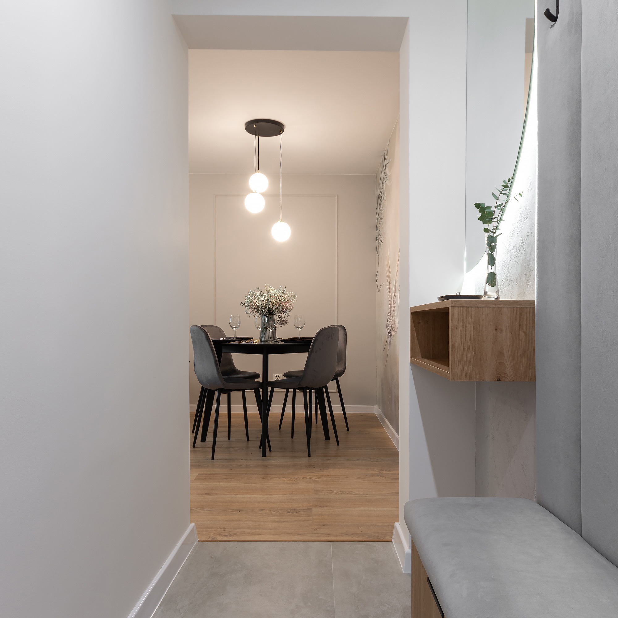 Wnętrze mieszkania z widokiem na stół jadalny z szarymi krzesłami, nakryty do posiłku, oświetlony wiszącą lampą z trzema kulami, z boku w przedpokoju wbudowana ławka z drewnianą półką pod lustrem.