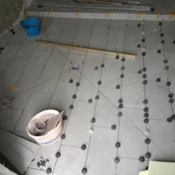 Szare płytki podłogowe w trakcie układania, z użyciem systemu poziomowania płytek, widoczny poziomica, wiadro i fragmenty płytek.