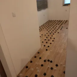 Parkiet laminowany z systemem poziomowania, widoczne klipsy i kliny, ściany przygotowane do malowania, okno zasłonięte roletą.