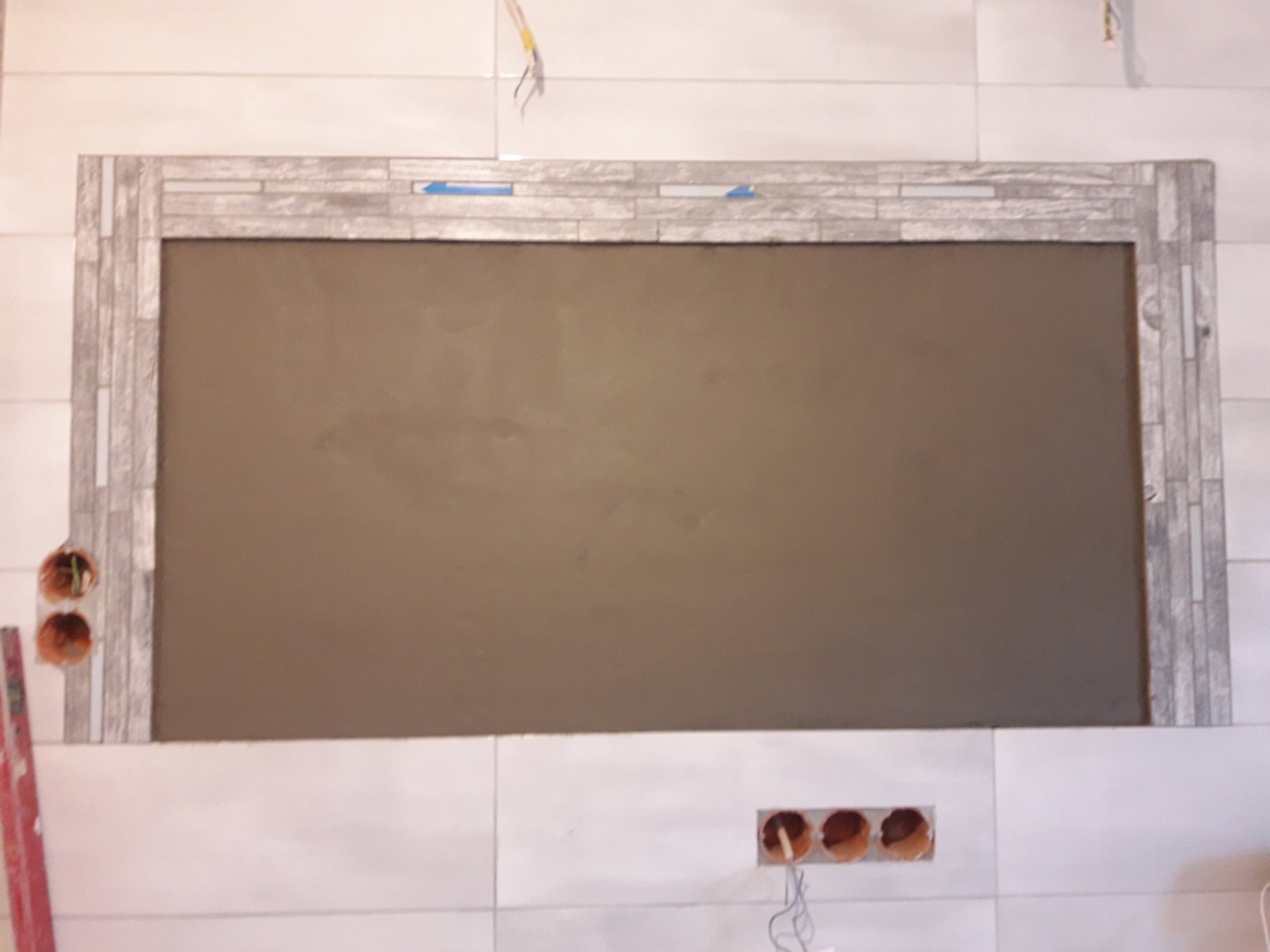 Ściana z białych płytek, z przygotowanym miejscem na lustro obłożonym dekoracyjną mozaiką w odcieniach szarości, widoczne puszki elektryczne.