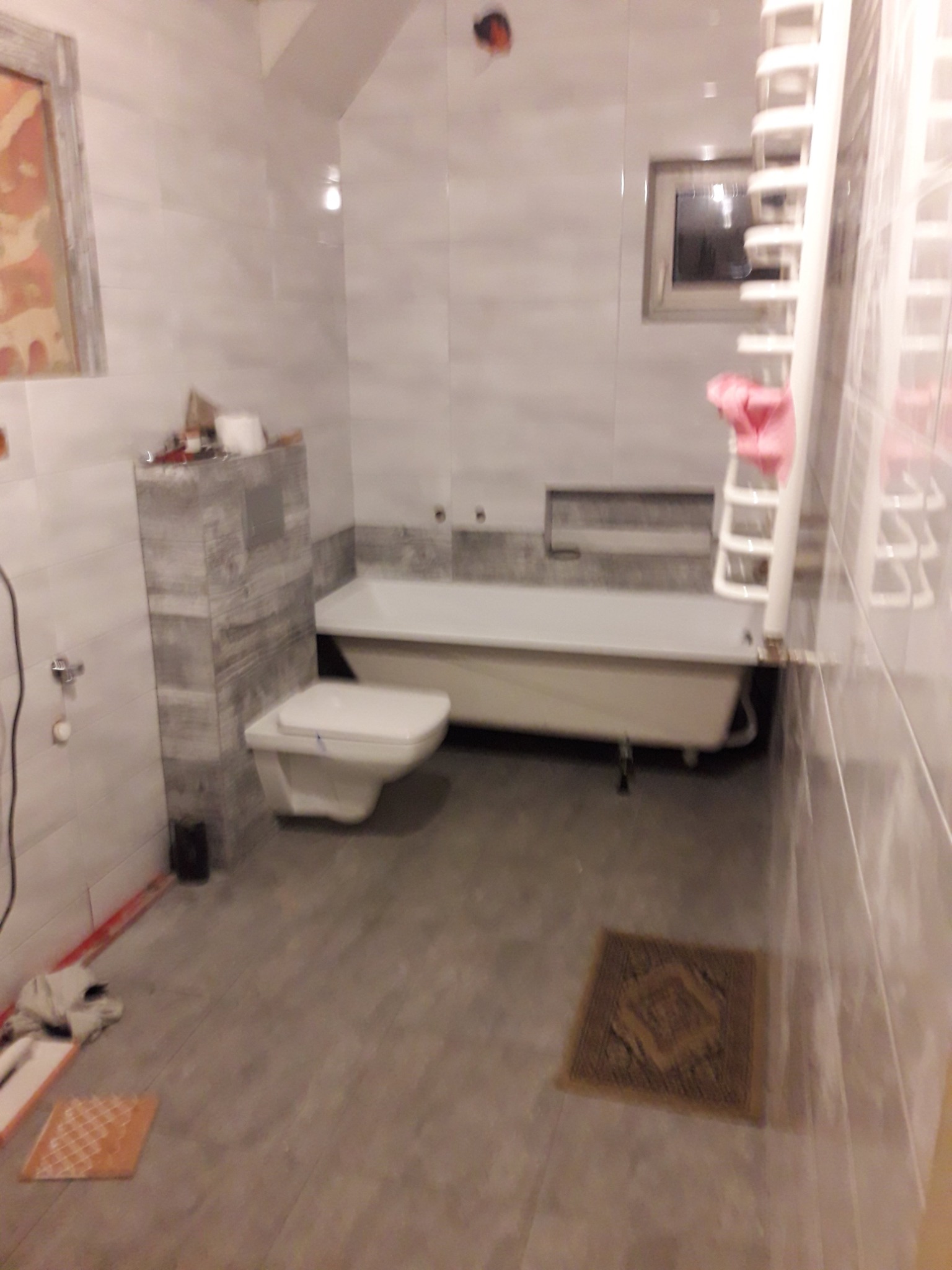 Wnętrze łazienki w trakcie prac: biała wanna, toaleta podwieszana na stelażu obłożonym płytkami imitującymi kamień, grzejnik drabinkowy, widoczne narzędzia i płytki na podłodze.