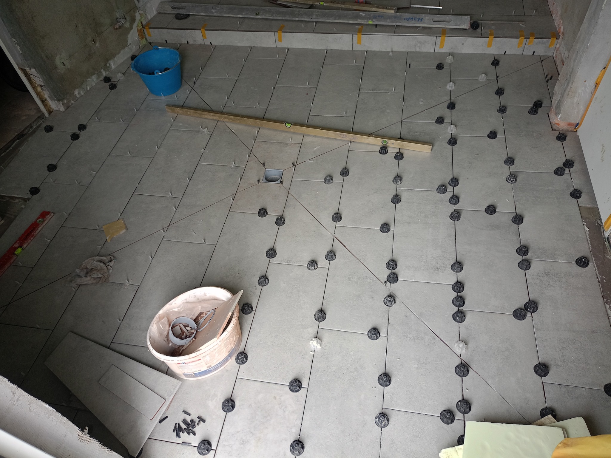 Szare płytki podłogowe w trakcie układania, z użyciem systemu poziomowania płytek, widoczny poziomica, wiadro i fragmenty płytek.