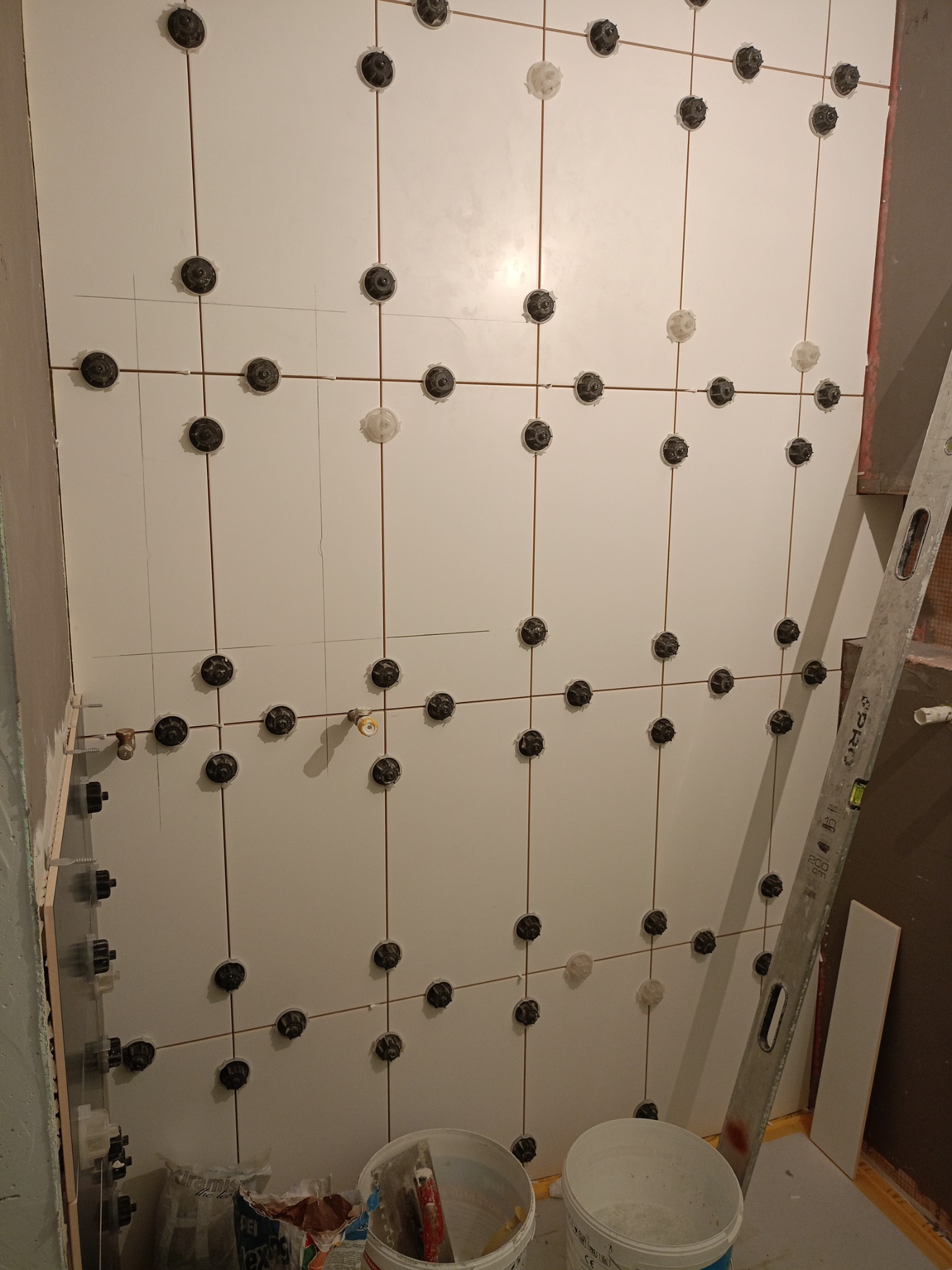 Świeżo ułożone białe płytki na ścianie z zastosowaniem systemu poziomowania płytek, widoczne czarne i białe klipsy oraz poziomica oparta o ścianę.