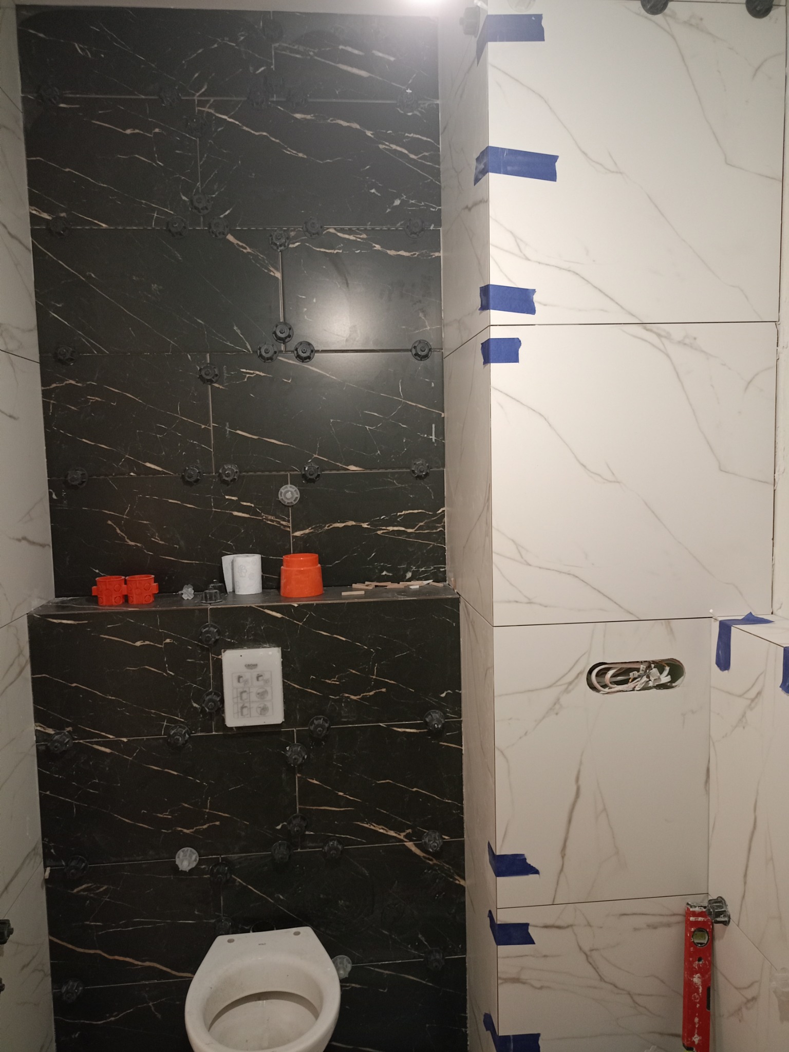 Remont łazienki: Czarno-białe płytki imitujące marmur na ścianach w trakcie układania, widoczne plastikowe krzyżyki dystansowe, poziomica i niepodłączona toaleta.