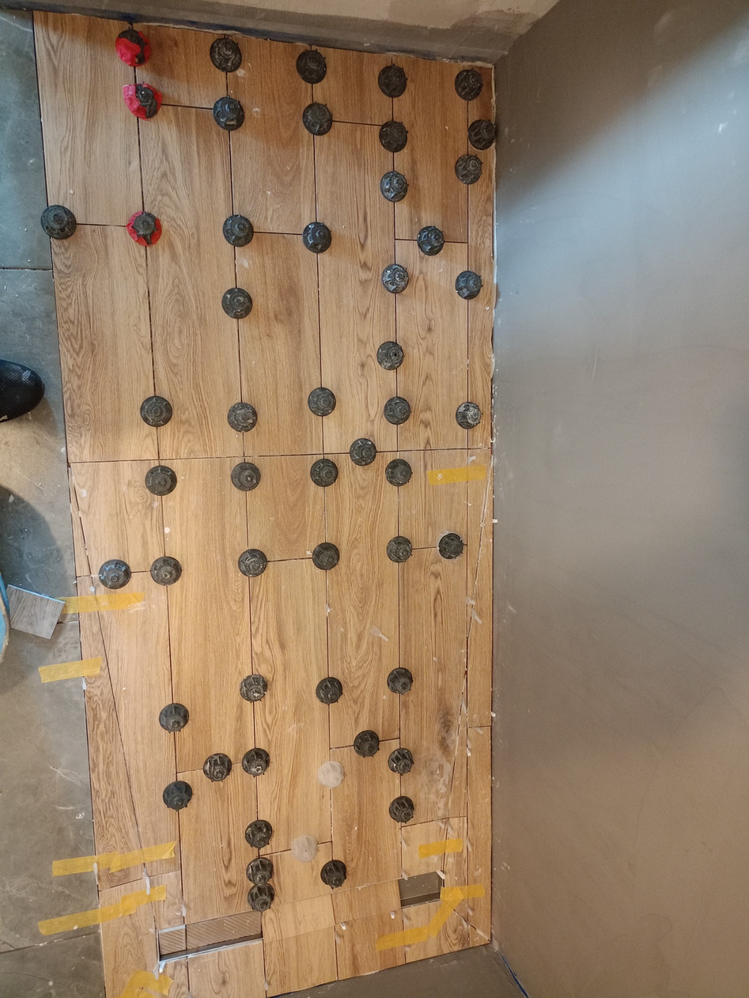 Układanie drewnopodobnych płytek podłogowych z użyciem systemu poziomowania, widoczne klipsy i kliny, fragment szarej ściany w tle.