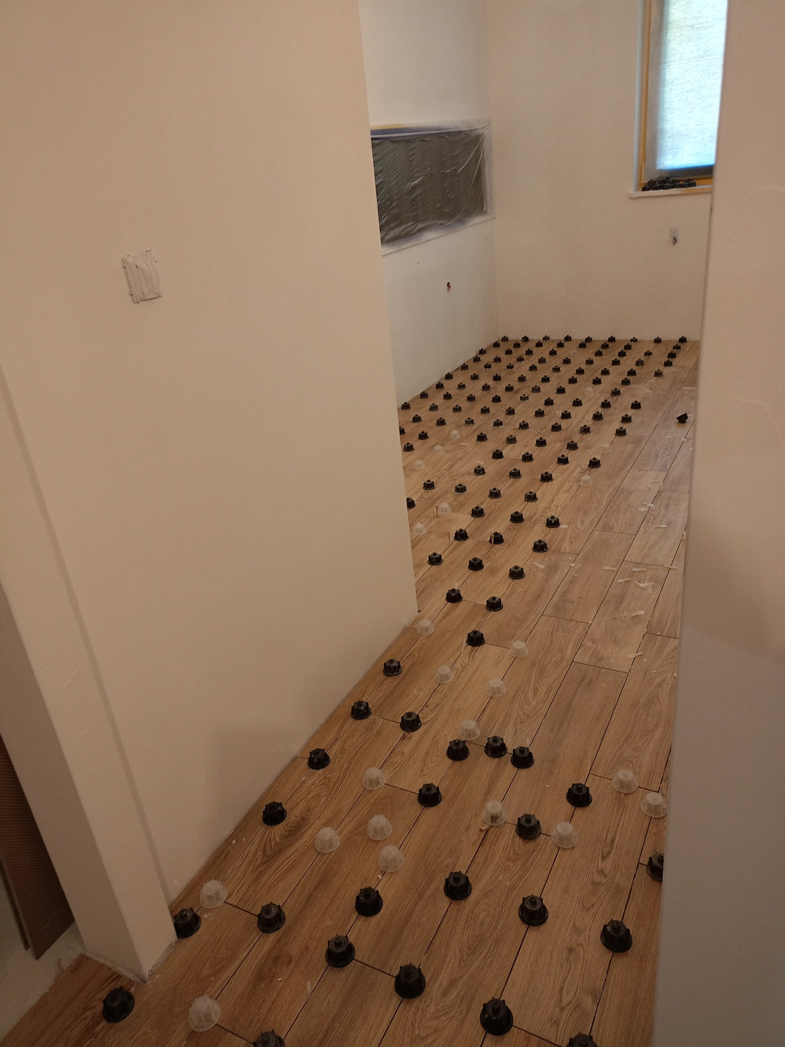 Parkiet laminowany z systemem poziomowania, widoczne klipsy i kliny, ściany przygotowane do malowania, okno zasłonięte roletą.