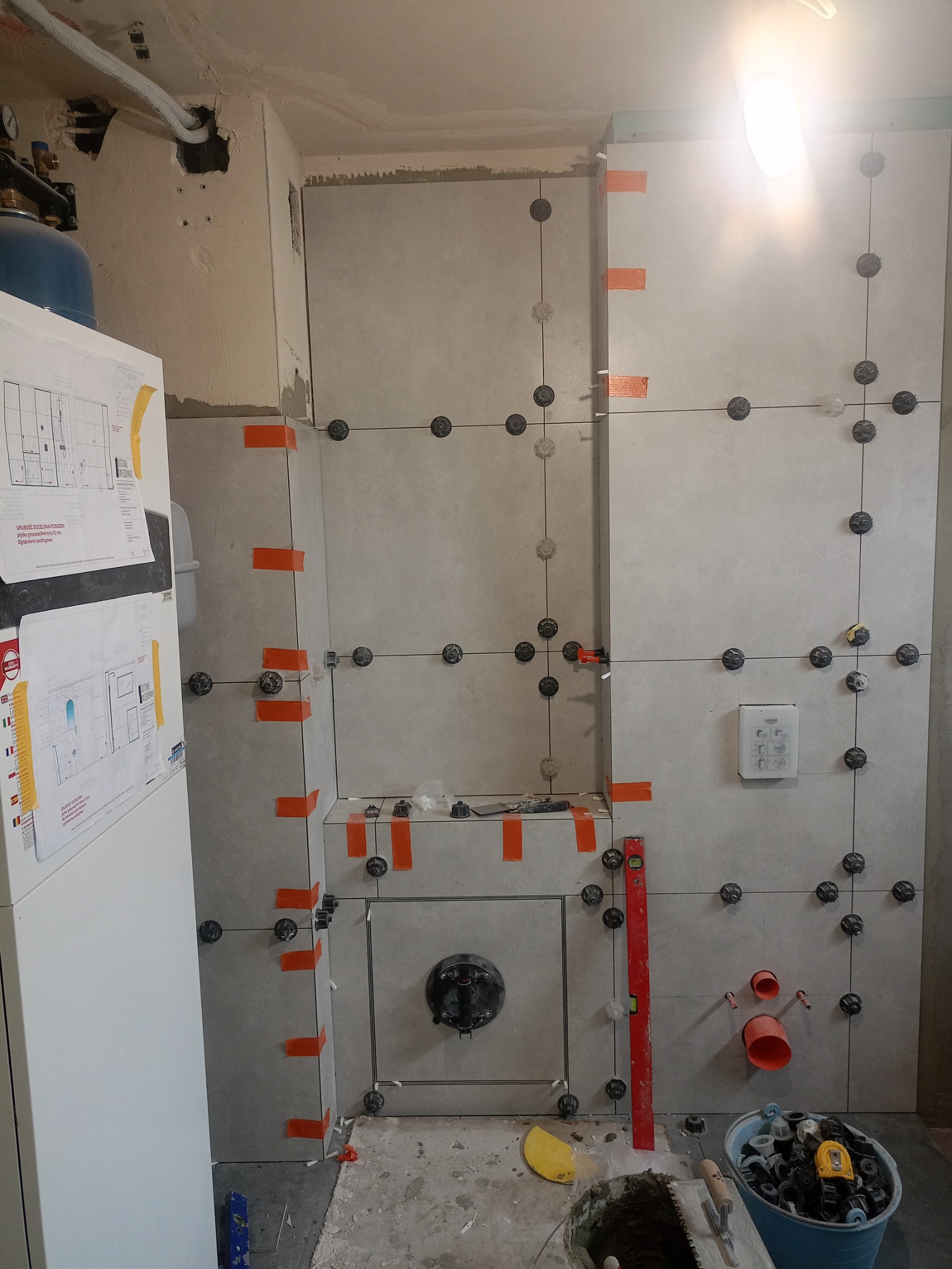 Ściana łazienki w trakcie układania płytek ceramicznych, widoczne przyssawki do płytek, poziomica, rury, wiadro z narzędziami i zaprawa, na ścianie przyklejone rysunki techniczne.