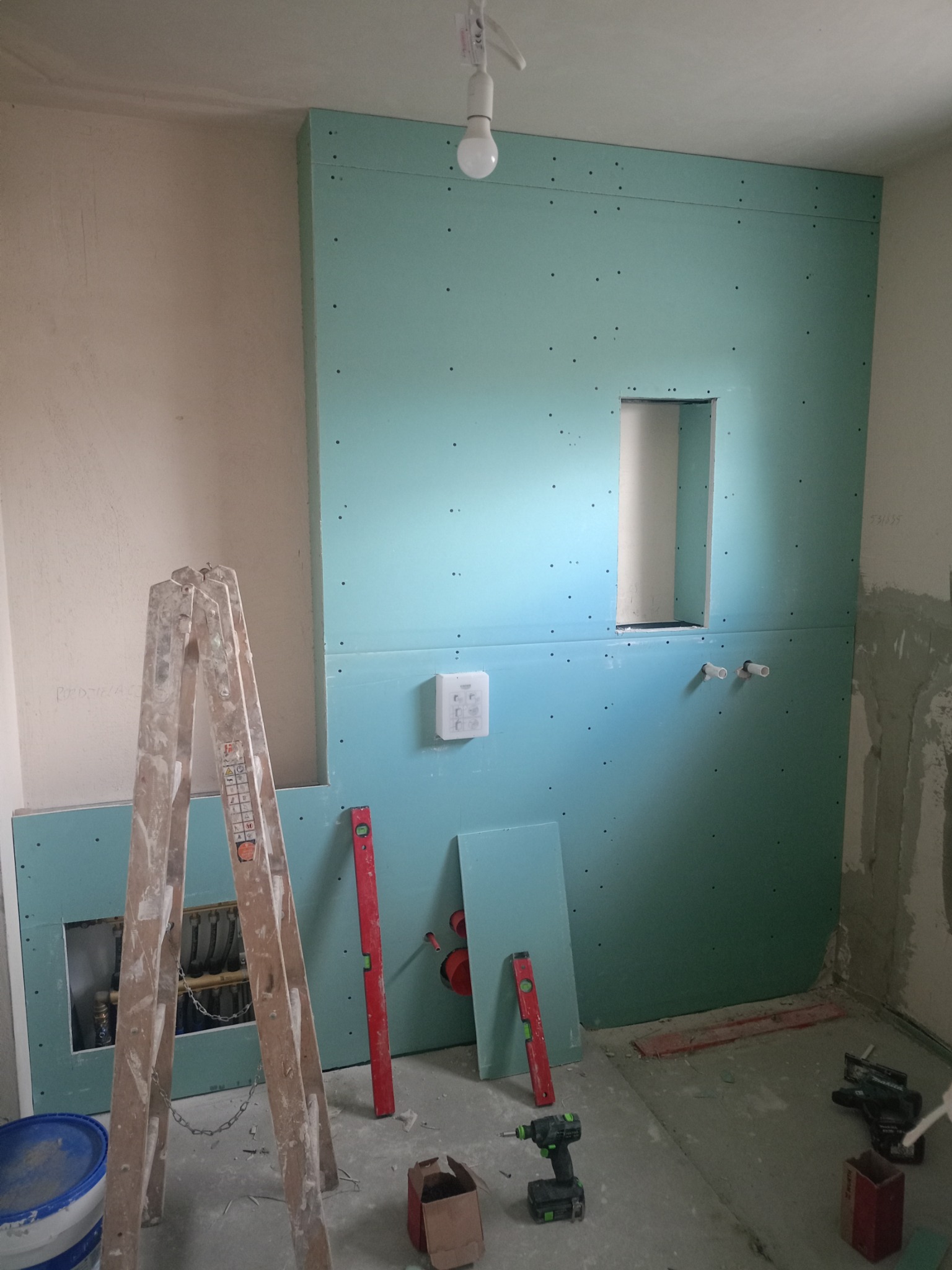 Montaż zielonej płyty karton gips w łazience w Pępowie, widoczne narzędzia, drabina i instalacja wodna w otworze.