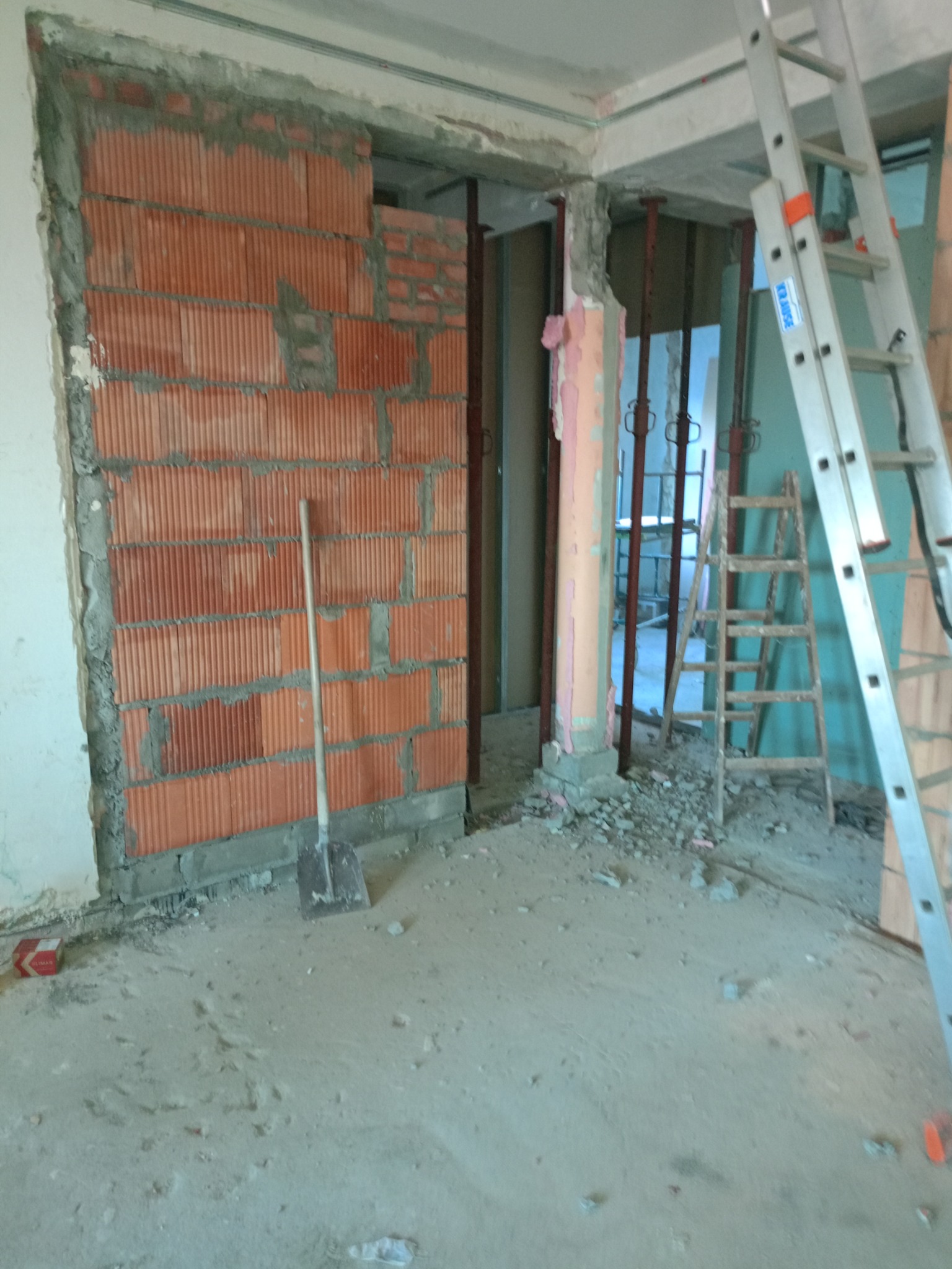 Wnętrze w trakcie prac murarskich: częściowo wymurowana ściana z czerwonej cegły kratówki, podparta metalowymi stemplami, obok drabina aluminiowa i drewniana, na podłodze gruz i szpadel.