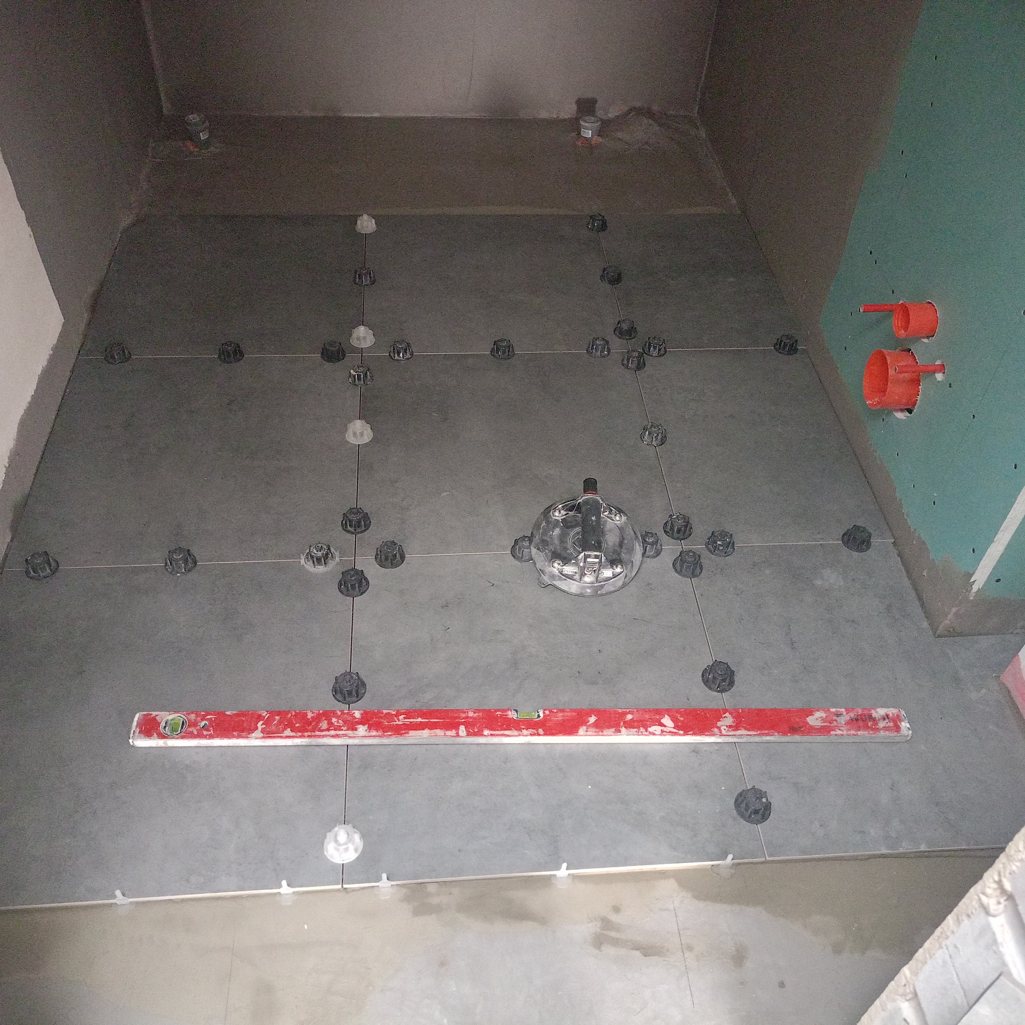 Szare płytki podłogowe w trakcie układania, z systemem poziomowania płytek, widoczna poziomica i otwory na rury w ścianie z płyt gipsowo-kartonowych.