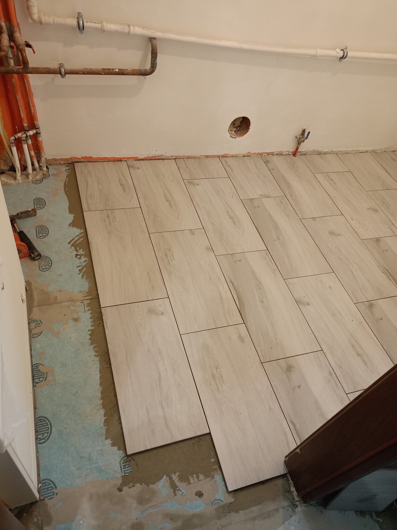 Fragment podłogi w trakcie układania jasnych, podłużnych płytek imitujących drewno, widoczna warstwa hydroizolacji Gardex oraz wystające rury instalacji wodnej