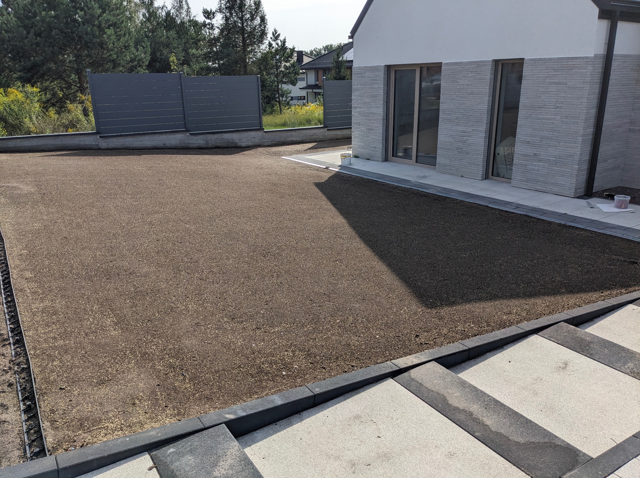 Przygotowany teren pod przyszły trawnik z widocznym systemem drenażowym wzdłuż ogrodzenia, obok nowoczesnego domu z szarymi elementami elewacji i tarasu.