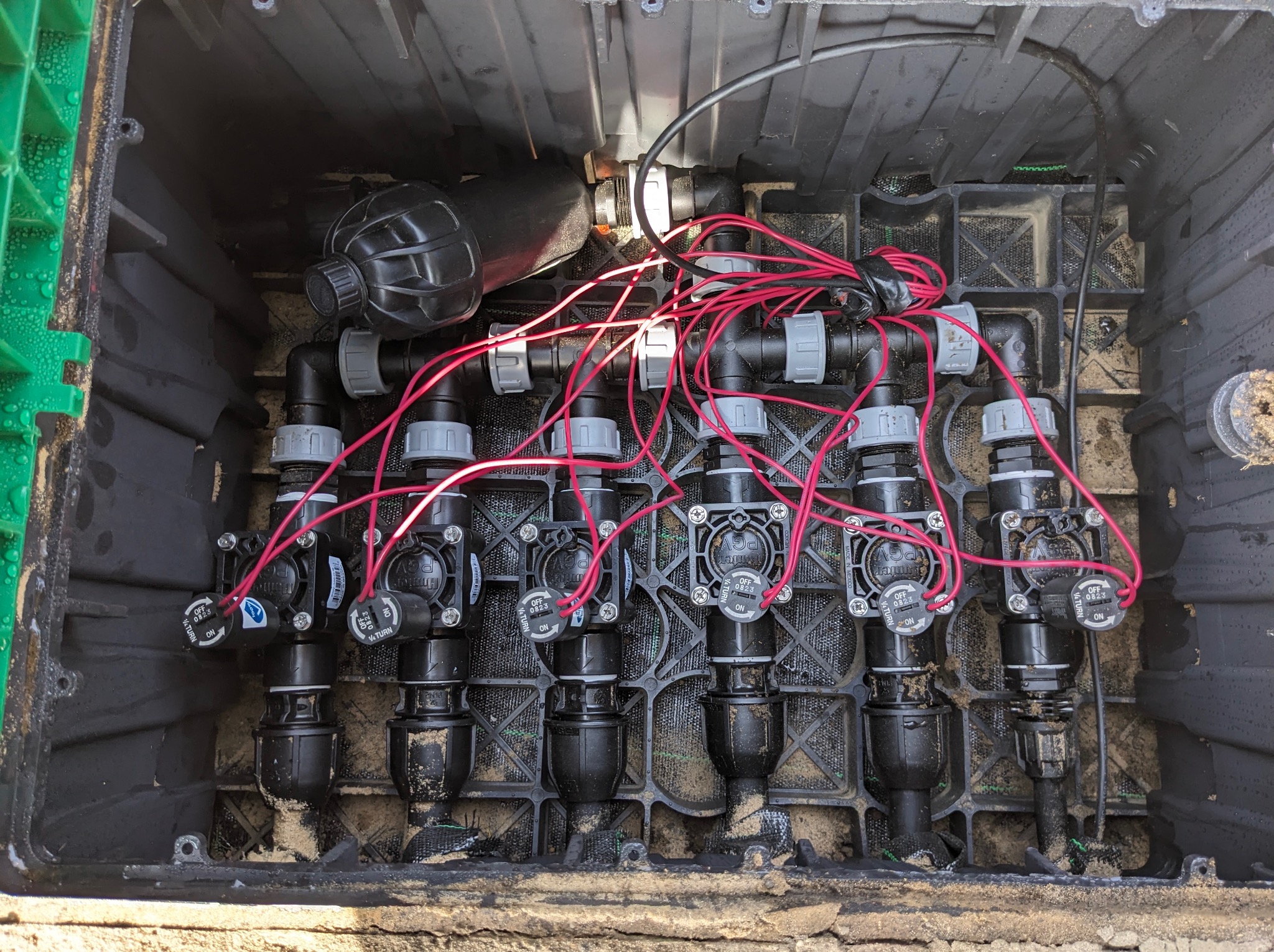 Skrzynka z zaworami elektromagnetycznymi i przewodami systemu nawadniania, widoczne napisy na zaworach i zabrudzenia piaskiem.