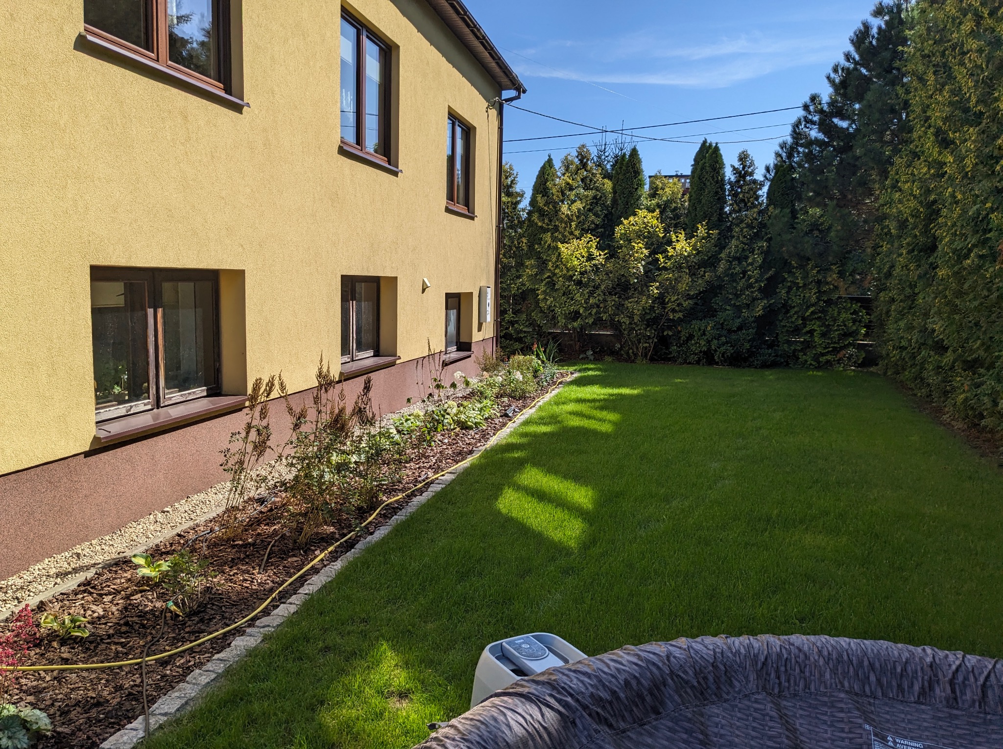 Widok na zadbany trawnik przydomowy z systemem nawadniania, obrzeża z kostki, rabata z korą i roślinami pod ścianą domu o żółtej elewacji, w tle wysokie tuje.