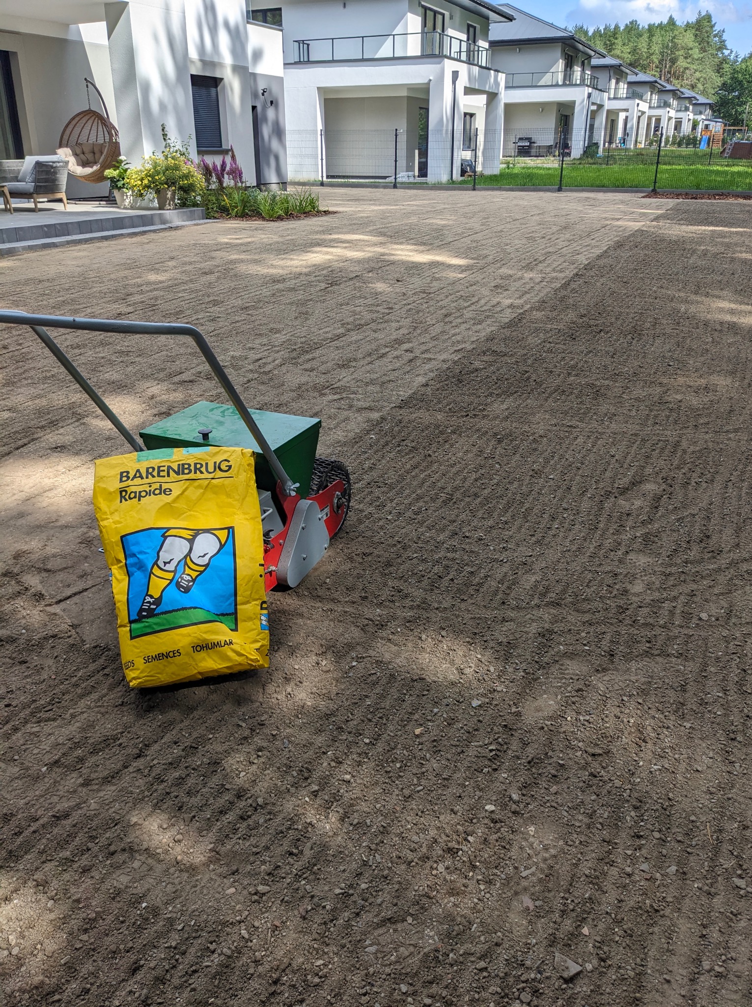Przygotowane podłoże z widocznymi śladami po siewniku, obok stoi siewnik i torba z nasionami trawy Barenbrug Rapide, w tle budynki mieszkalne.