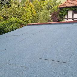 HORUS-DACH - Pracownik w trakcie układania papy termozgrzewalnej na płaskim dachu; widoczny fragment dachu z nową, niebieską izolacją, narzędzia pracy i otoczenie z zielenią.