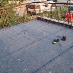 HORUS-DACH - Płaski dach pokryty papą, z widocznymi łączeniami, na którym leżą robocze rękawice; w tle metalowa balustrada, zieleń i maszyna budowlana.