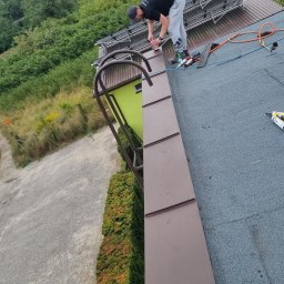 HORUS-DACH - Pracownik w czarnej koszulce i szarych spodniach na dachu pokrytym papą, zabezpiecza obróbkę blacharską przy użyciu silikonu dekarskiego; w tle drabina dekarska i zielone drzewa.