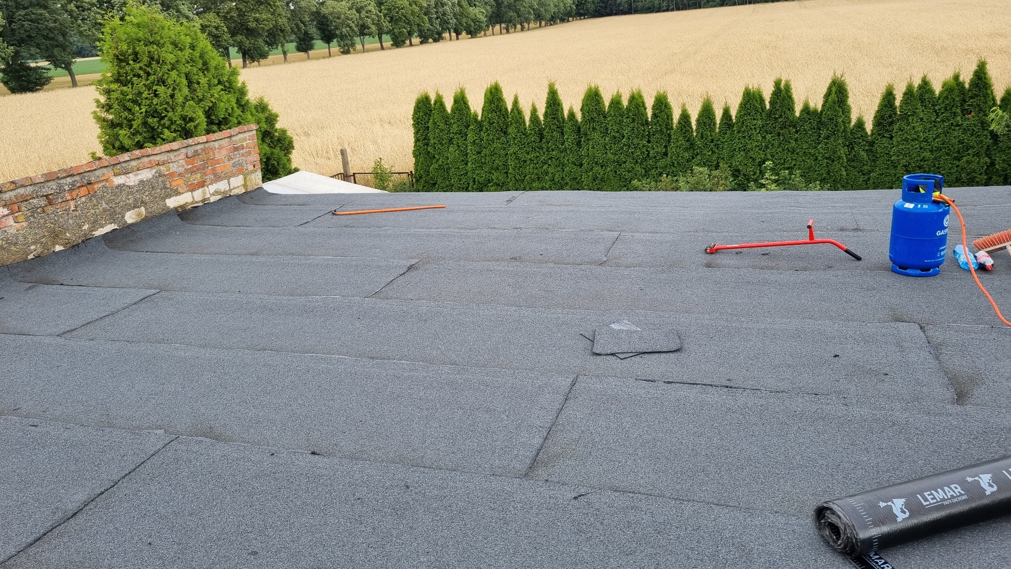 Dach pokryty papą termozgrzewalną, widoczny fragment muru, butla gazowa, narzędzia dekarskie i rolka papy LEMAR na tle pola i rzędu tuj.