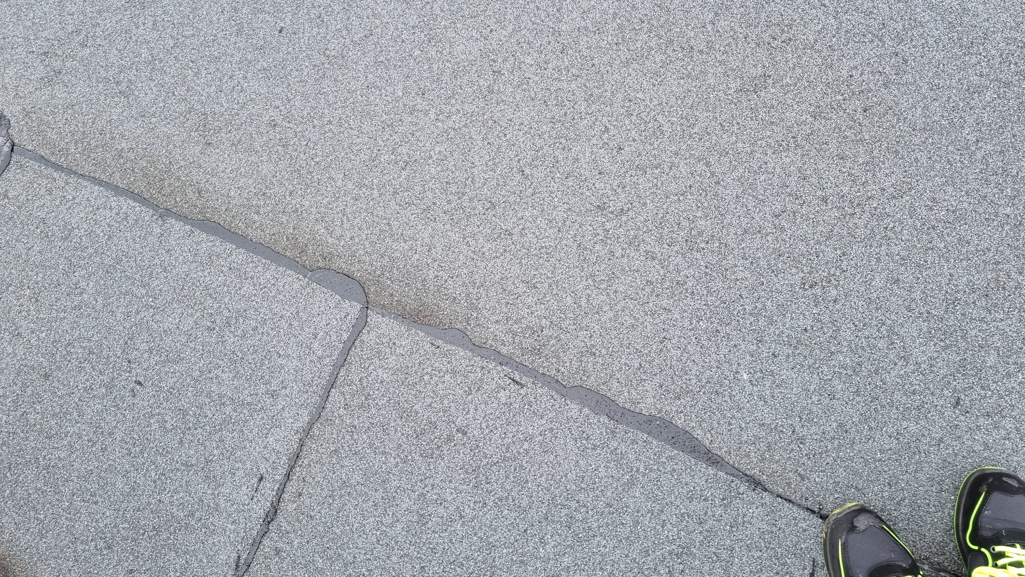 Szczegółowe ujęcie szarego pokrycia dachowego z widocznymi łączeniami płyt i uszczelnieniami, częściowo mokrymi, z fragmentem butów sportowych w prawym dolnym rogu.