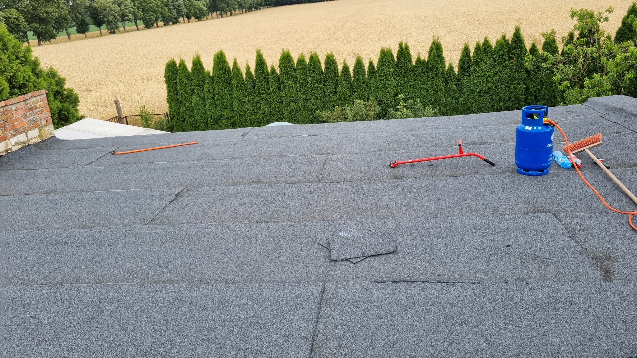 Szary dach pokryty papą termozgrzewalną z widocznym sprzętem dekarskim: butla gazowa, szczotka, narzędzie do zgrzewania, z tłem zielonych drzew i pola.