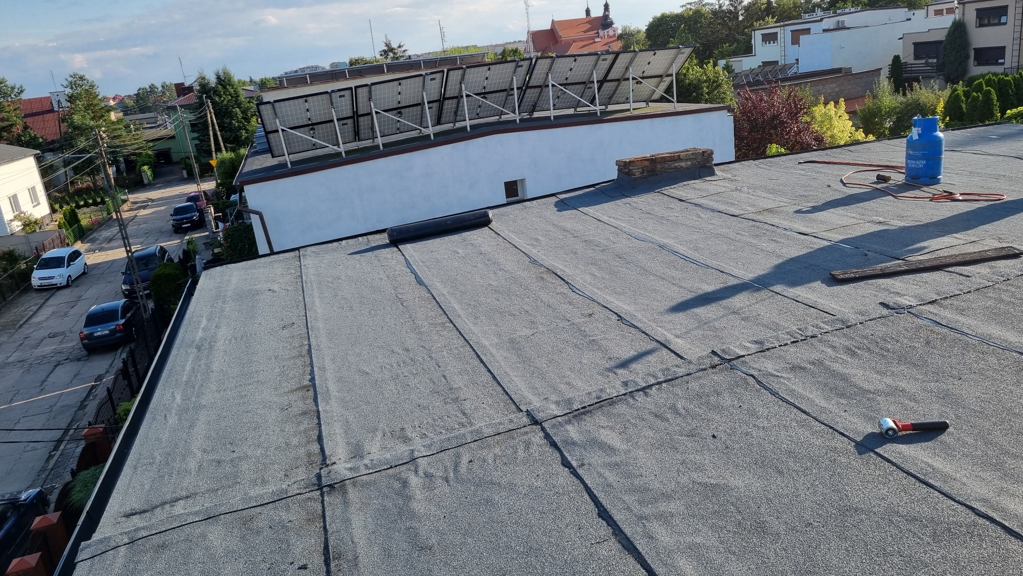 Szary dach pokryty papą termozgrzewalną z widocznym butlą gazową, palnikiem dekarskim, wałkiem dociskowym i niezrolowanym fragmentem papy, panele słoneczne na sąsiednim dachu, zabudowa miejska w tle.